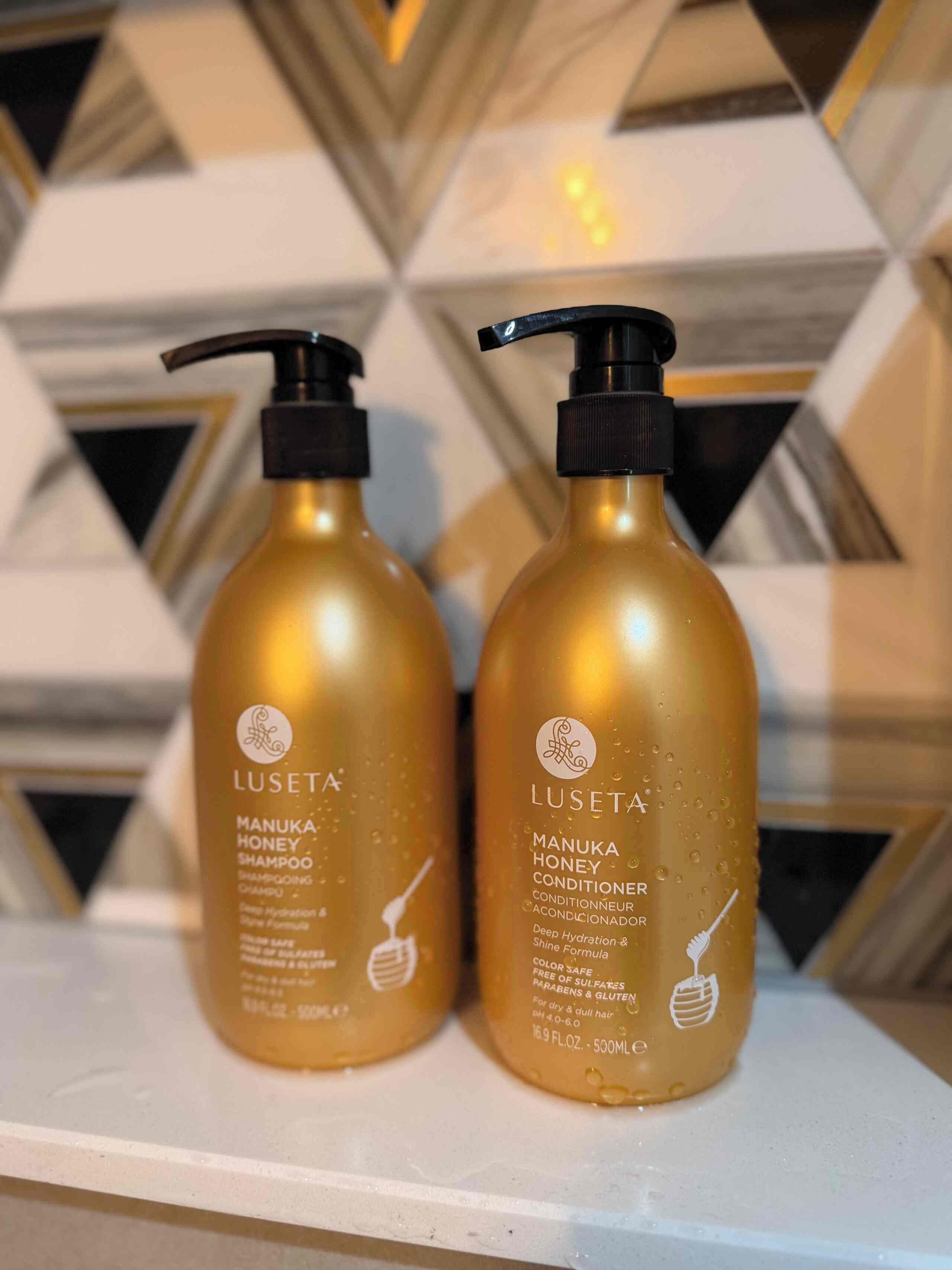 Dos botellas de champú y acondicionador de miel de Manuka Lusetta sobre una superficie blanca con un fondo de diseño geométrico.