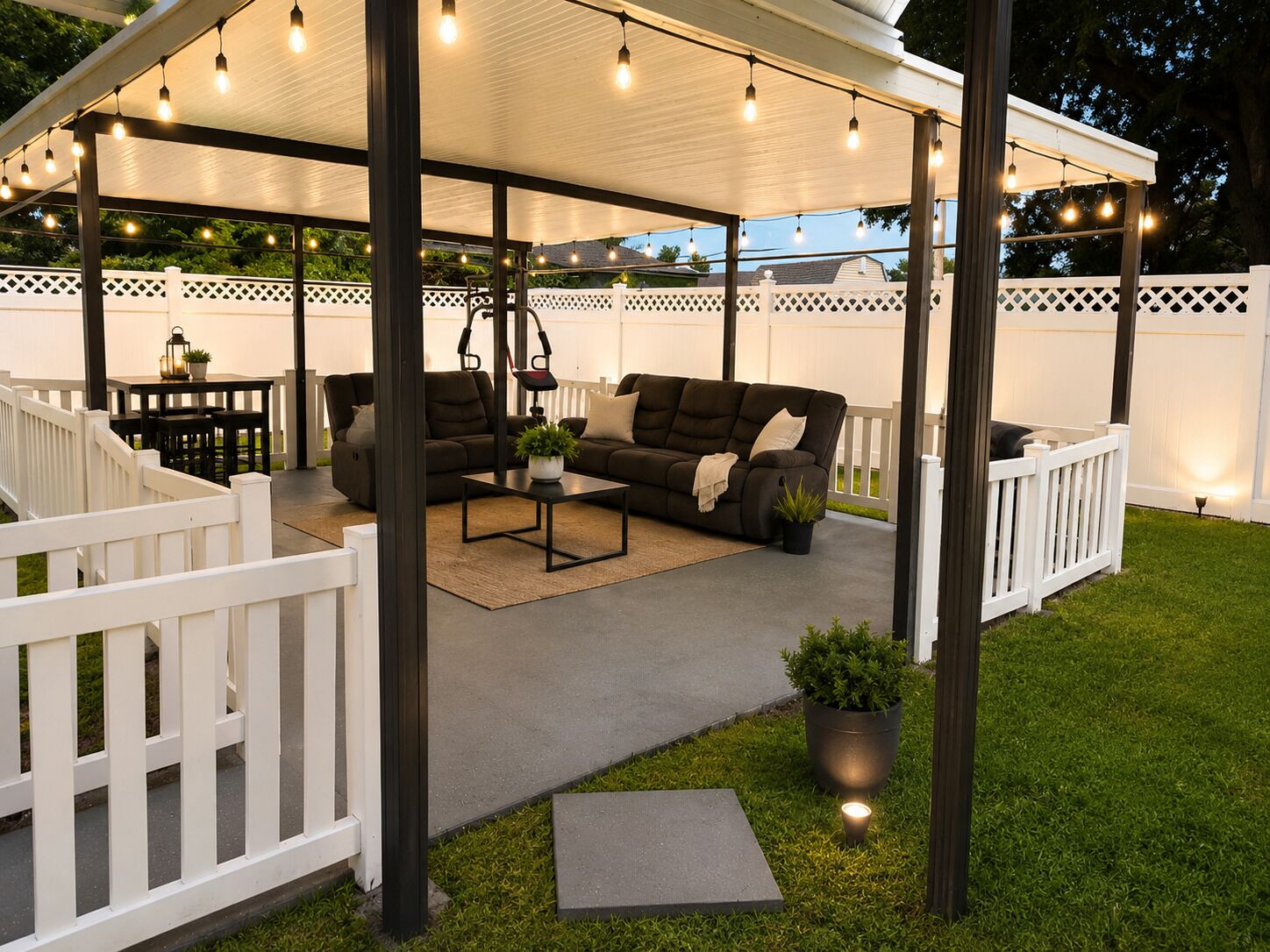 A covered outdoor seating area with a sofa, table, and chairs, surrounded by a white fence.