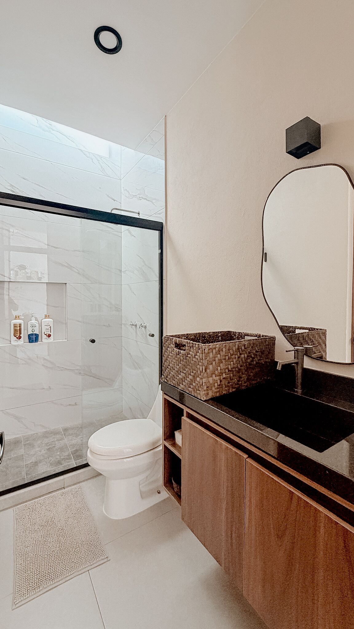 Un baño con cabina de ducha de vidrio, inodoro blanco, tocador de madera con espejo y un estante pequeño con artículos de aseo.