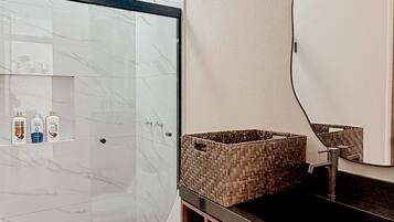 Un baño con cabina de ducha de vidrio, inodoro blanco, tocador de madera con espejo y un estante pequeño con artículos de aseo.