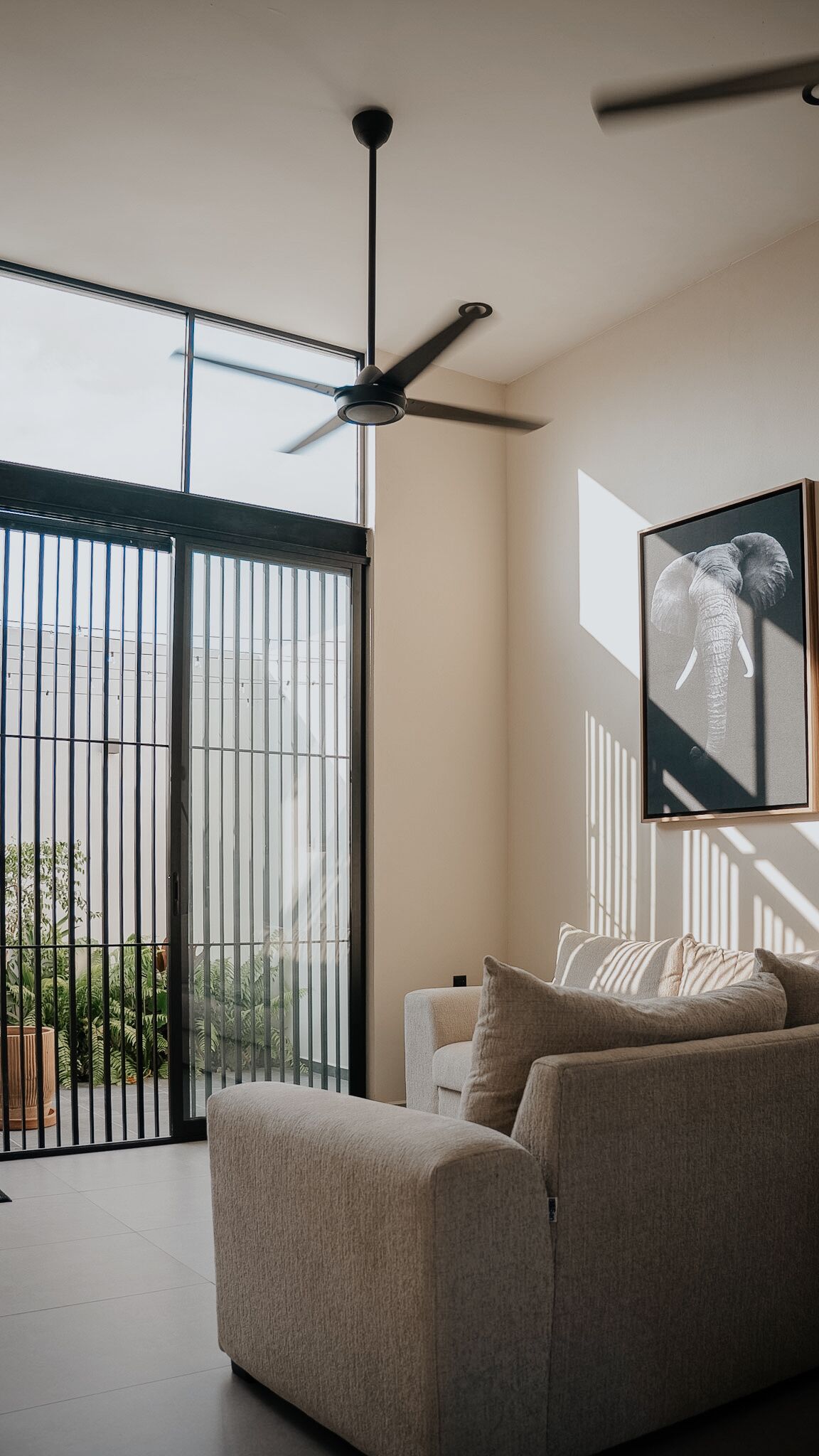 Un salon moderne avec un canapé, un ventilateur de plafond et une grande fenêtre équipée de stores verticaux.