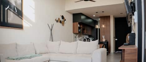 A modern living room with a sectional sofa, a ceiling fan, and a kitchen area in the background.
