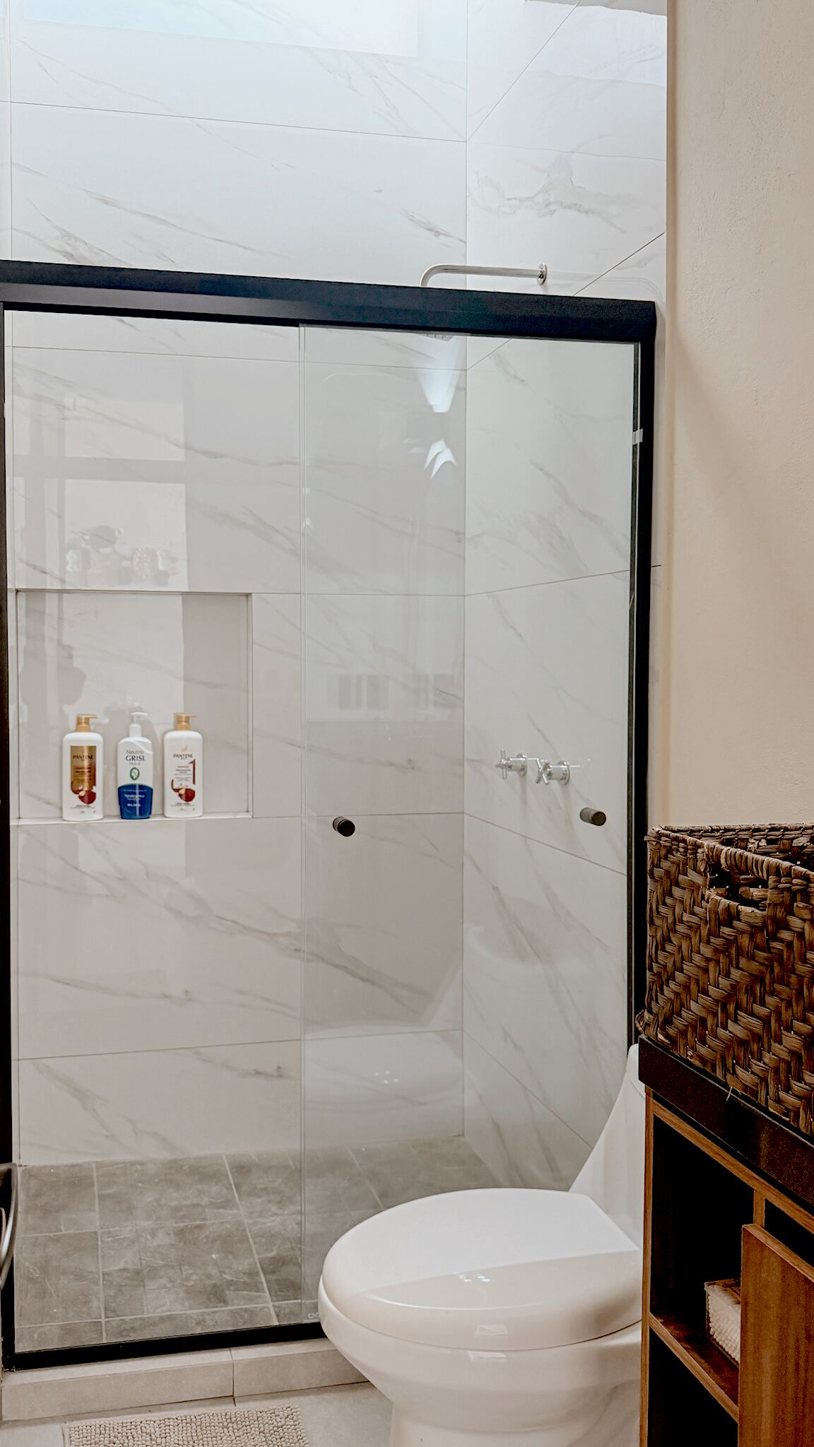 Un baño moderno con ducha de vidrio, inodoro blanco y repisa con artículos de aseo.