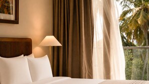 A hotel room with a bed, a headboard, a lamp, and a view of palm trees through sheer curtains.