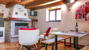 A rustic interior with a stone fireplace, wooden ceiling beams, a dining table with chairs, and a television mounted on the wall. - Rustico Grace by Interhome (Malvaglia)