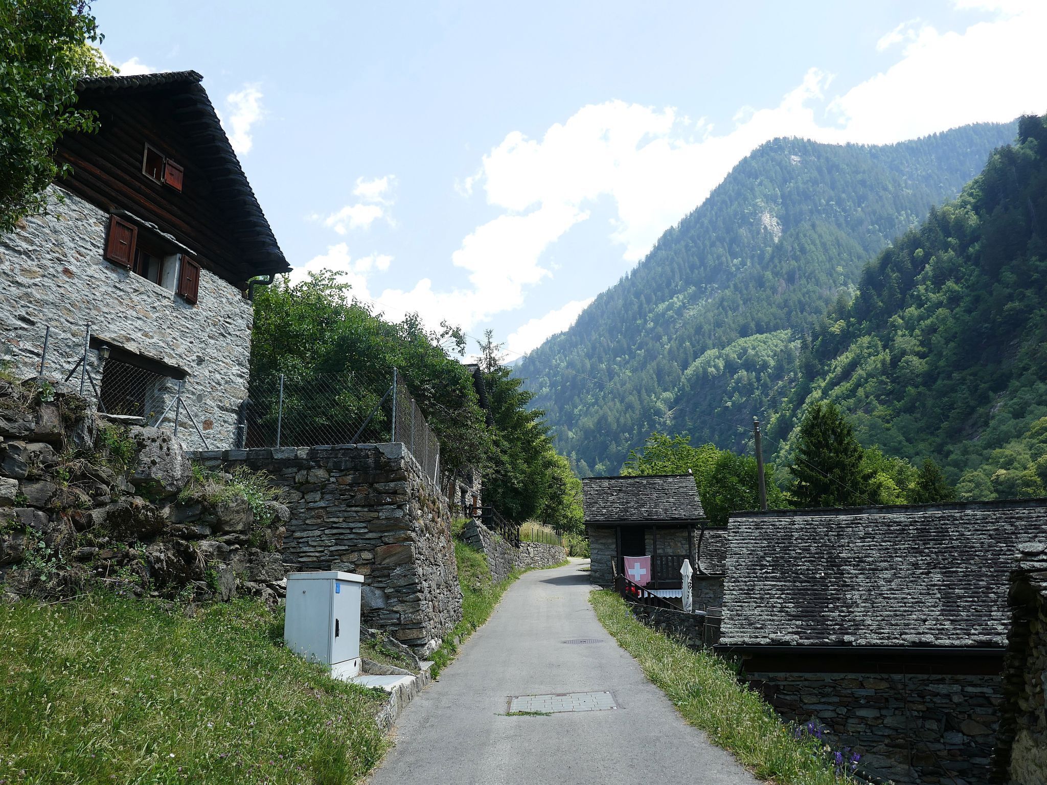 A narrow rural road flanked by stone buildings with a mountainous backdrop.