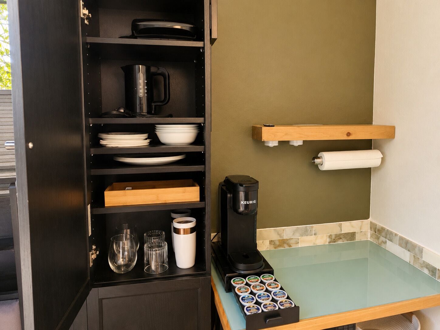 A kitchen with a coffee maker, shelves with plates and bowls, and a glass-top counter with a coffee pod holder.