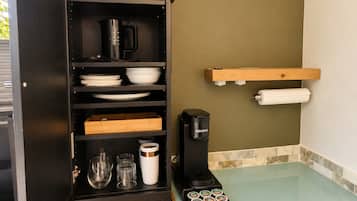 A kitchen with a coffee maker, shelves with plates and bowls, and a glass-top counter with a coffee pod holder.