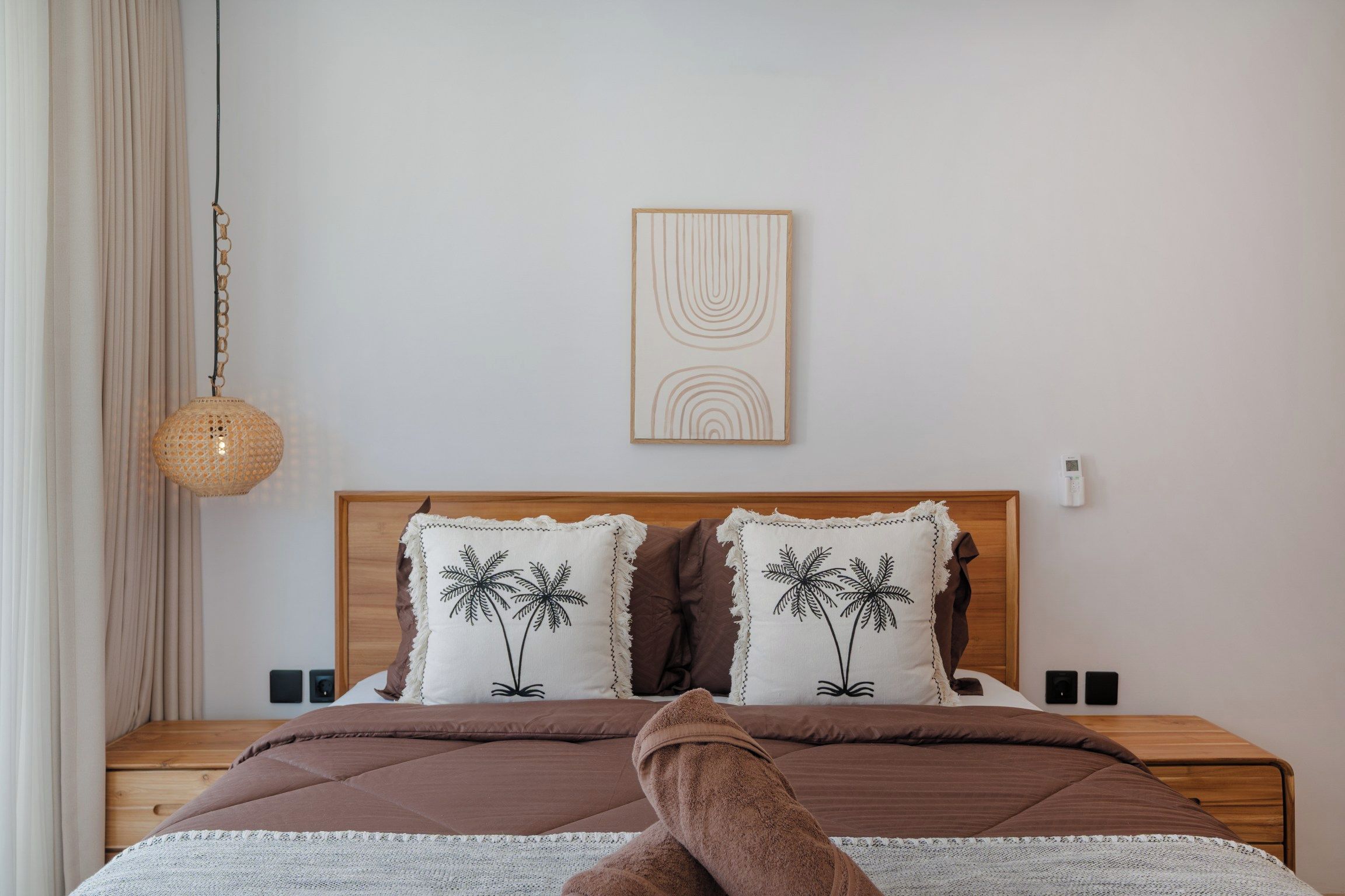 Ein Schlafzimmer mit einem hölzernen Kopfende, zwei Kissen mit Palmenmotiven, einem gewebten Pendel und einem Nachttisch mit Wandthermostat.