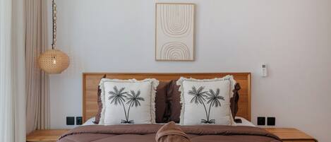 Une chambre avec une tête de lit en bois, deux oreillers ornés de motifs de palmiers, un luminaire suspendu en fibres tressées et une table de chevet dotée d’un thermostat fixé au mur.
