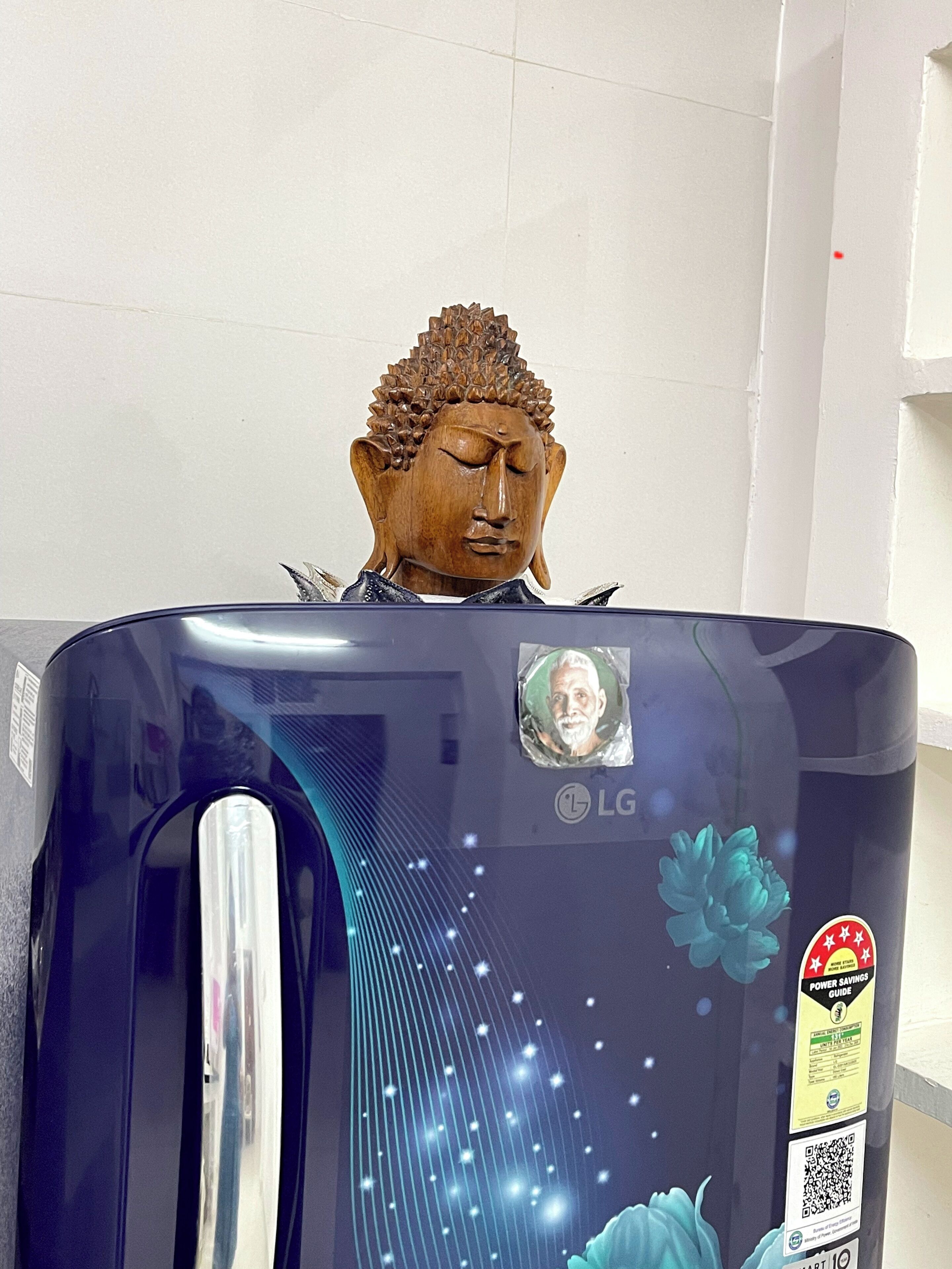 A refrigerator with a Buddha head sculpture on top, a sticker with a QR code, and a power savings guide sticker.