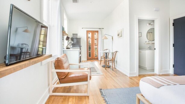 A modern, well-lit room with a large window, a flat-screen TV, a leather chair, a small dining table, and a bed.