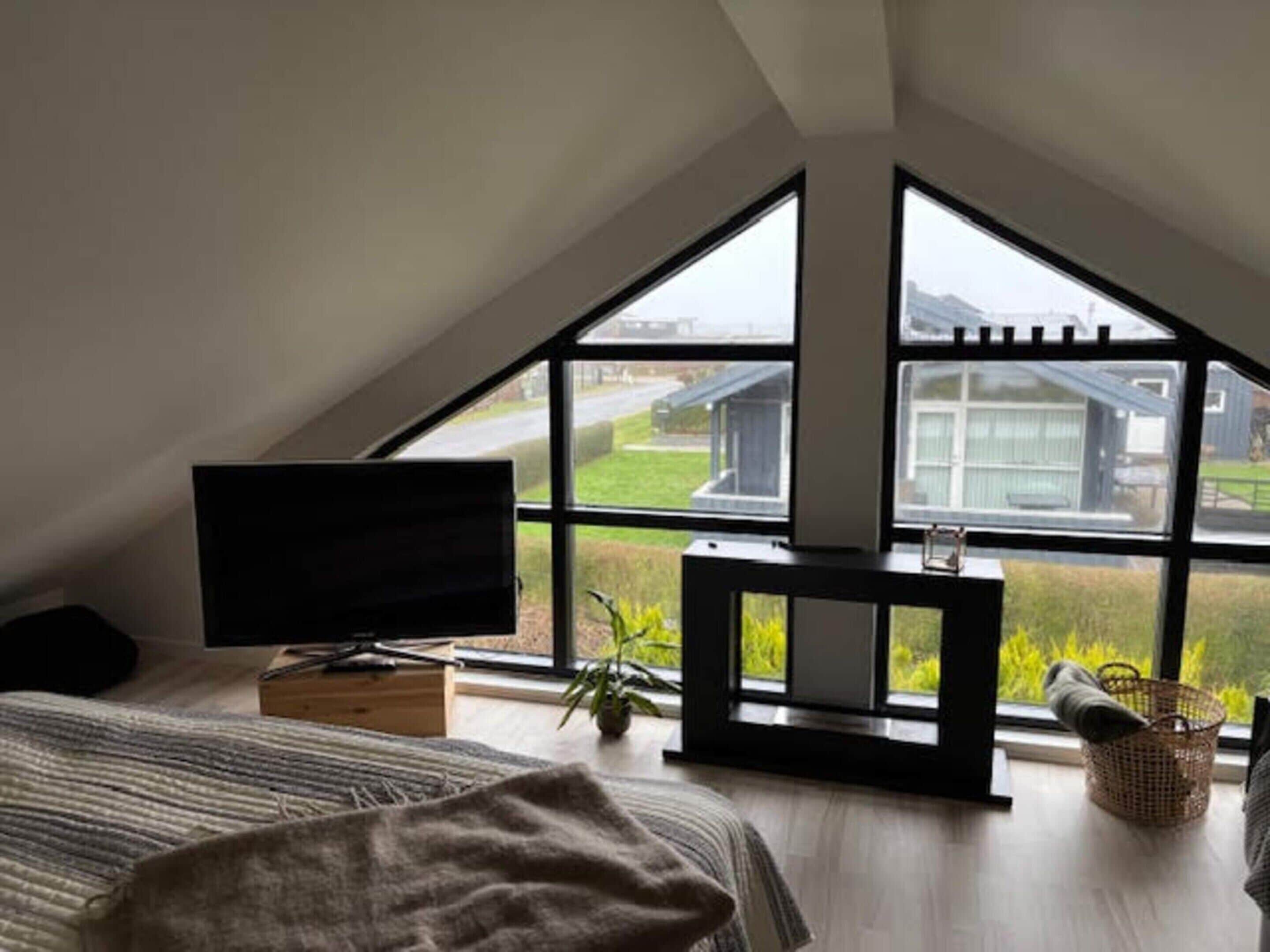A bedroom with a sloped ceiling, a television on a stand, a bed with a striped blanket, a small table with a plant, and a window with a view of a house and greenery.