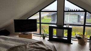 Ett sovrum med ett sluttande tak, en tv på ett stativ, en säng med ett randigt överkast, ett litet bord med en växt och ett fönster med utsikt över ett hus och grönska.