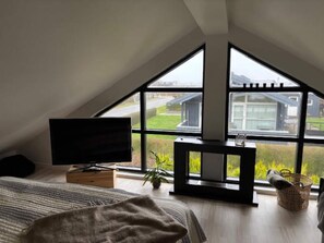A bedroom with a sloped ceiling, a television on a stand, a bed with a striped blanket, a small table with a plant, and a window with a view of a house and greenery. - 4 Star Holiday Home in Otterup (Otterup)