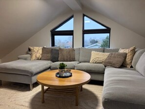 A modern loft bedroom with a sloped ceiling, large windows, a sectional sofa, a round wooden coffee table, and a small potted plant. - 4 Star Holiday Home in Otterup (Otterup)