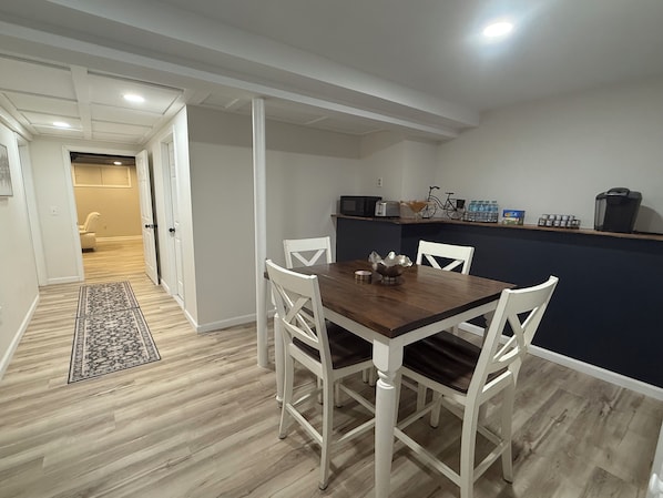 Een eetgedeelte met een houten tafel en witte stoelen, een keuken met apparatuur, en een gang die naar een andere kamer leidt.