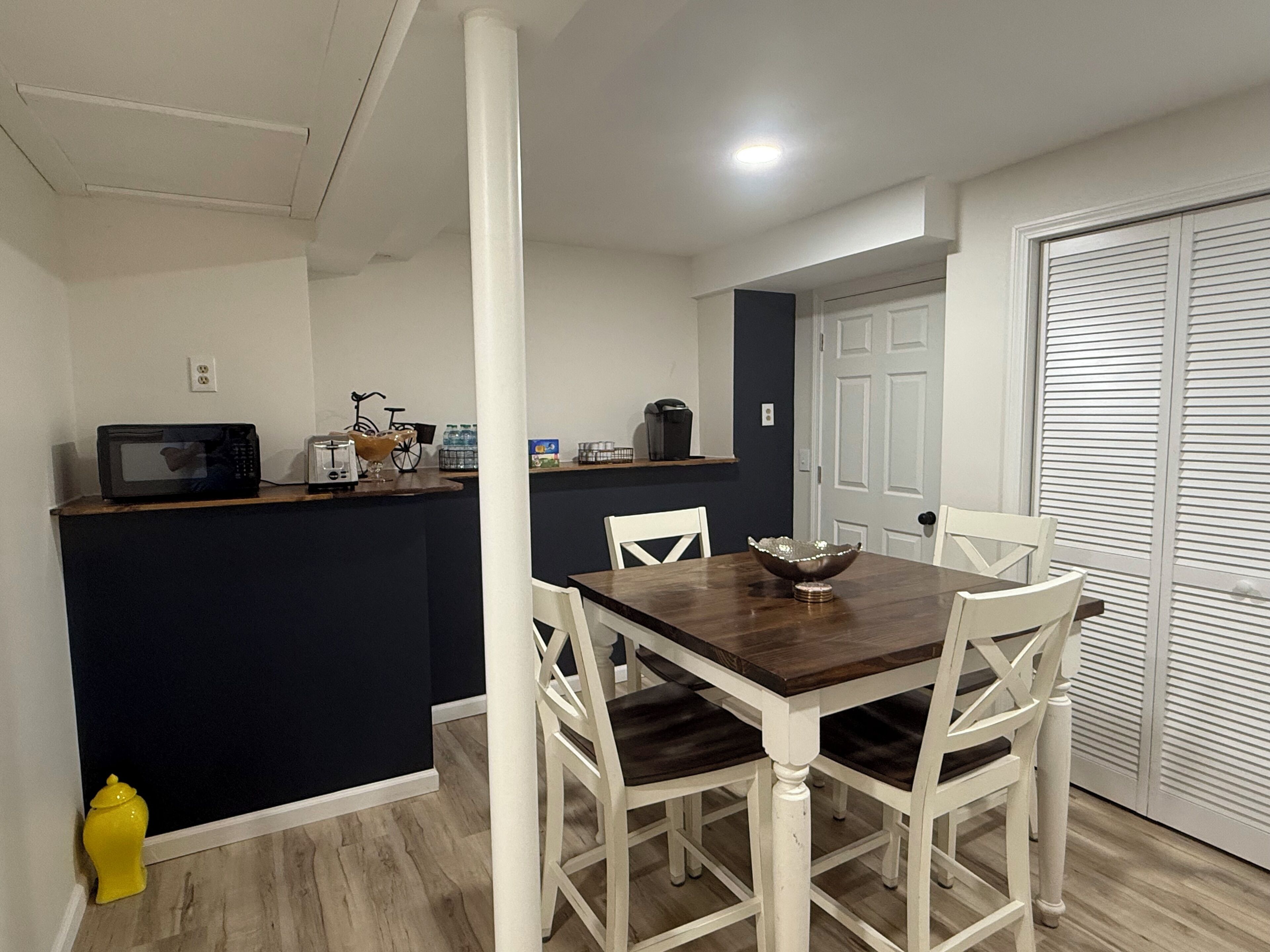 Un comedor con una mesa de madera y sillas blancas, un microondas en la encimera y una puerta con contraventanas.
