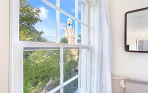 A room with a large window offering a view of a building with a tower, a white curtain, and a mirror on the wall. - Park House, Studio 3A on Monterey Square (Savannah)