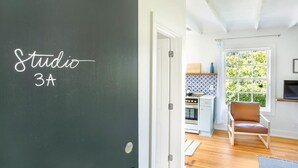 Huoneessa on tauluseinä, jossa lukee ”Studio 3A”, ja ikkunasta avautuu näkymä keittiöön, jossa on liesi ja tuoli.