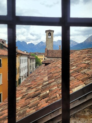 Vista pela janela de uma cidade com telhados de terracota, uma torre e montanhas ao fundo.