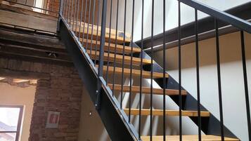 Escada de madeira com corrimão de metal, parede de tijolo e um ventilador em um ambiente com iluminação natural.