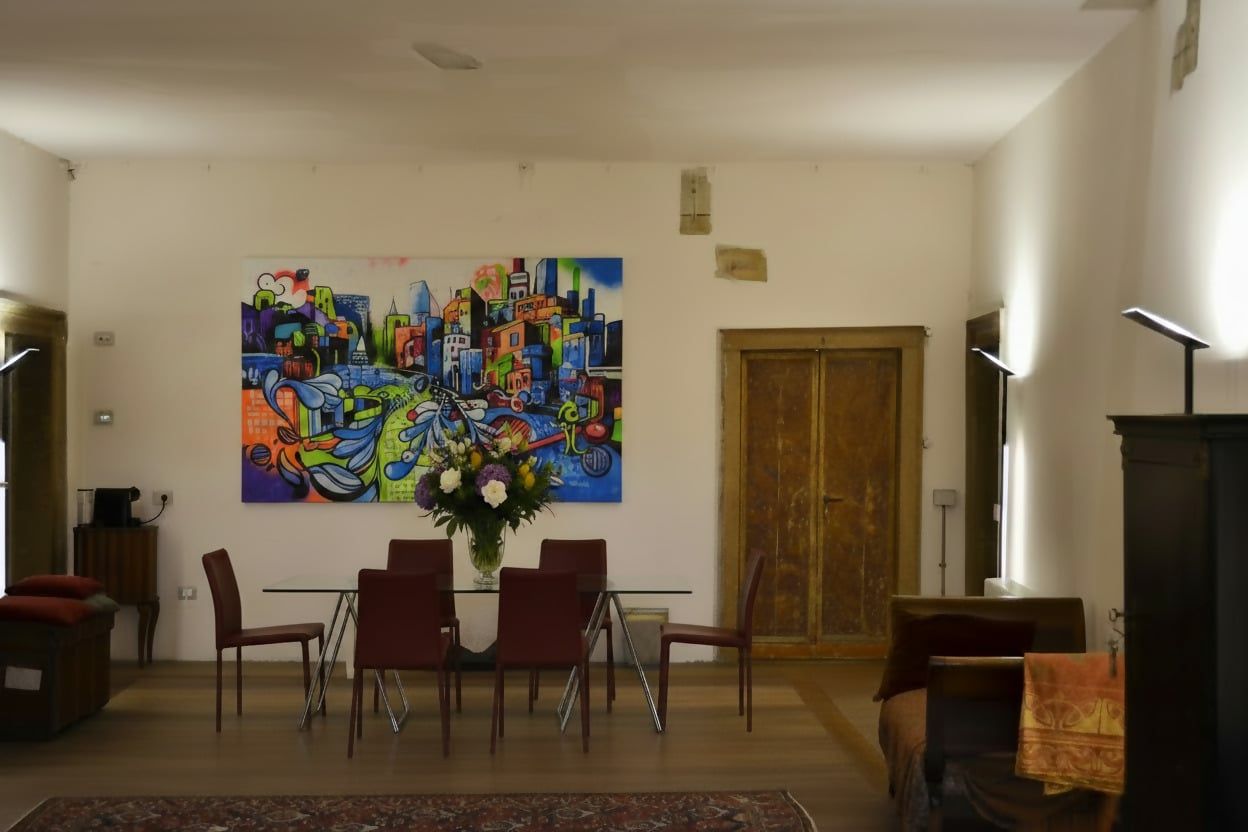 Uma sala de jantar com mesa e cadeiras, um quadro grande e colorido na parede, e uma porta de madeira.