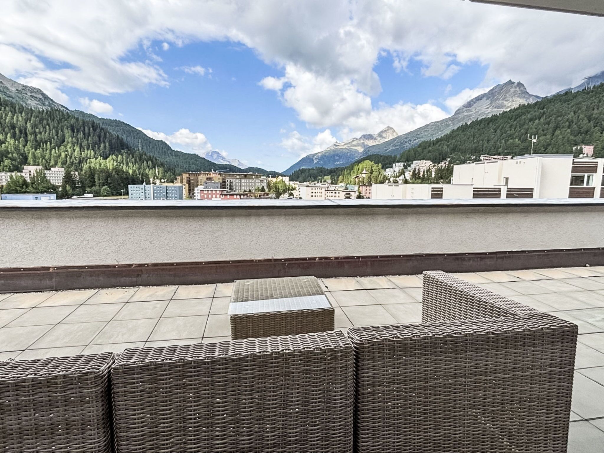A rooftop terrace with wicker furniture, overlooking a cityscape and mountains.