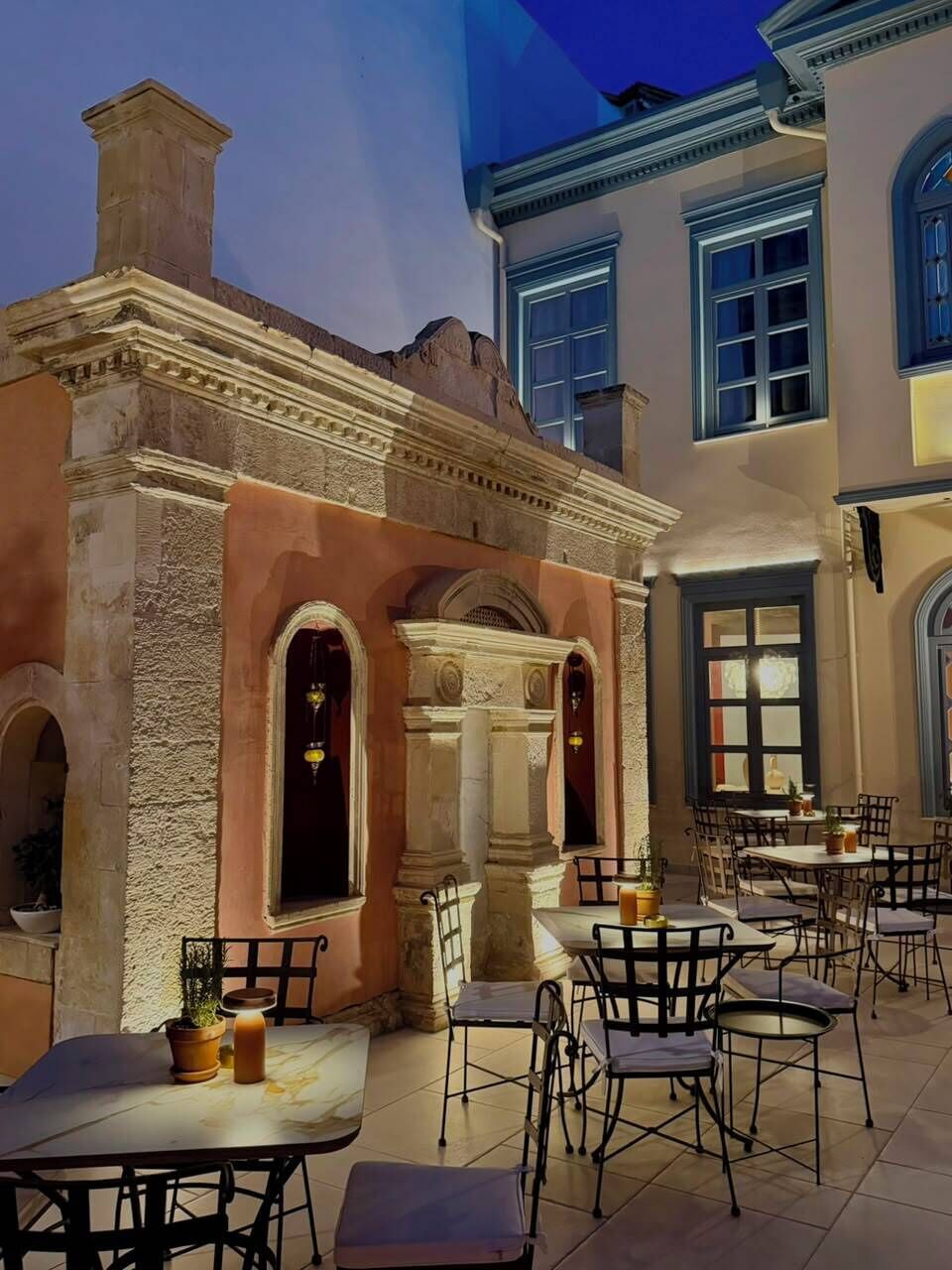 Un espacio de comedor al aire libre con varias mesas y sillas, un edificio de piedra con arcos y un amplio ventanal con vistas.