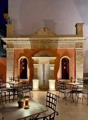 Un área de comedor al aire libre con mesas y sillas, un arco de piedra y una fuente.