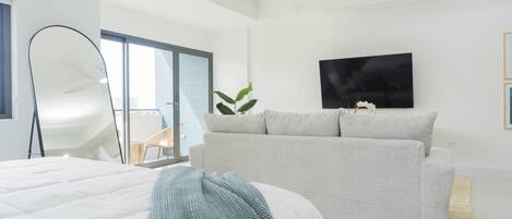 A modern bedroom with a large bed, a grey sofa, a flat-screen TV, and a balcony with a view.