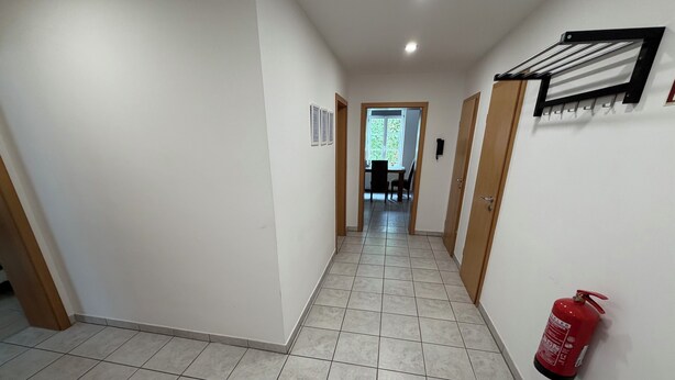 Um corredor com piso de azulejos, um extintor de incêndio fixado na parede e uma porta que dá para um quarto com janela.