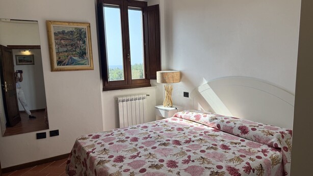 A bedroom with a bed, a window, a radiator, a lamp, and a painting on the wall. - Villa Le Cicale in Procchio (Marciana)