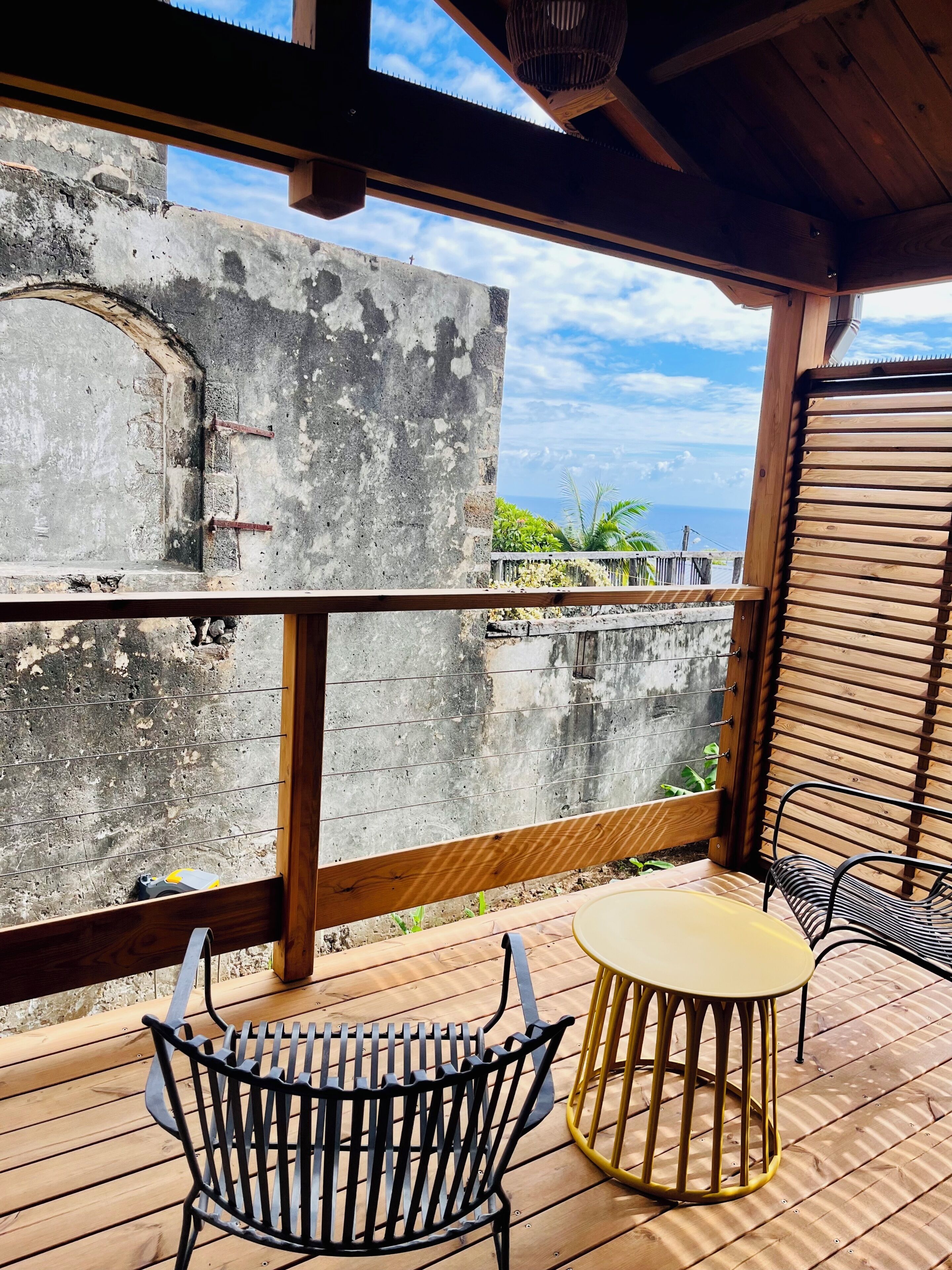 Um deck de madeira com mesa e cadeiras, com vista para a costa, mar e vegetação.