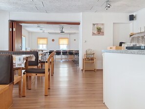 Una habitación amplia con piso de madera, un comedor con mesas y sillas, una cocina con mostrador y un pequeño rincón de descanso con una butaca.
