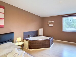 Un baño moderno con una bañera grande, una mesita de noche con lámpara y una ventana con vista a la vegetación.