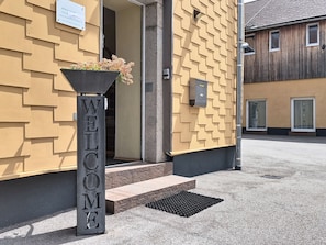 A building entrance with a 'WELCOME' sign, a planter with flowers, and a door leading inside.