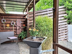 Een overdekt terras met een houten bankje, een plantenbak met bloemen en een pergola erboven.