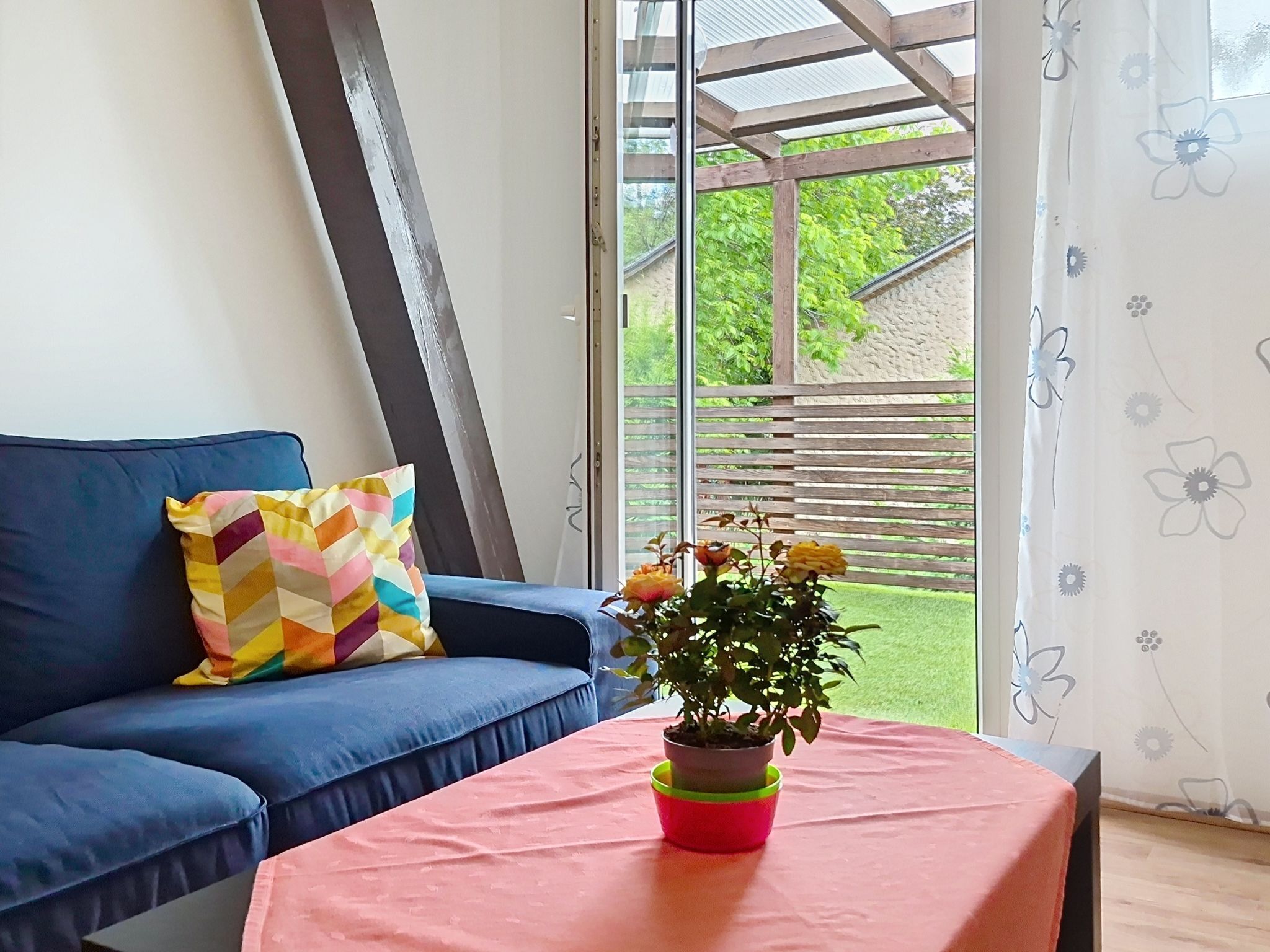Une pièce avec un canapé bleu, un coussin coloré, une petite table sur laquelle est posée une plante en pot, et une vue sur un jardin par la fenêtre.