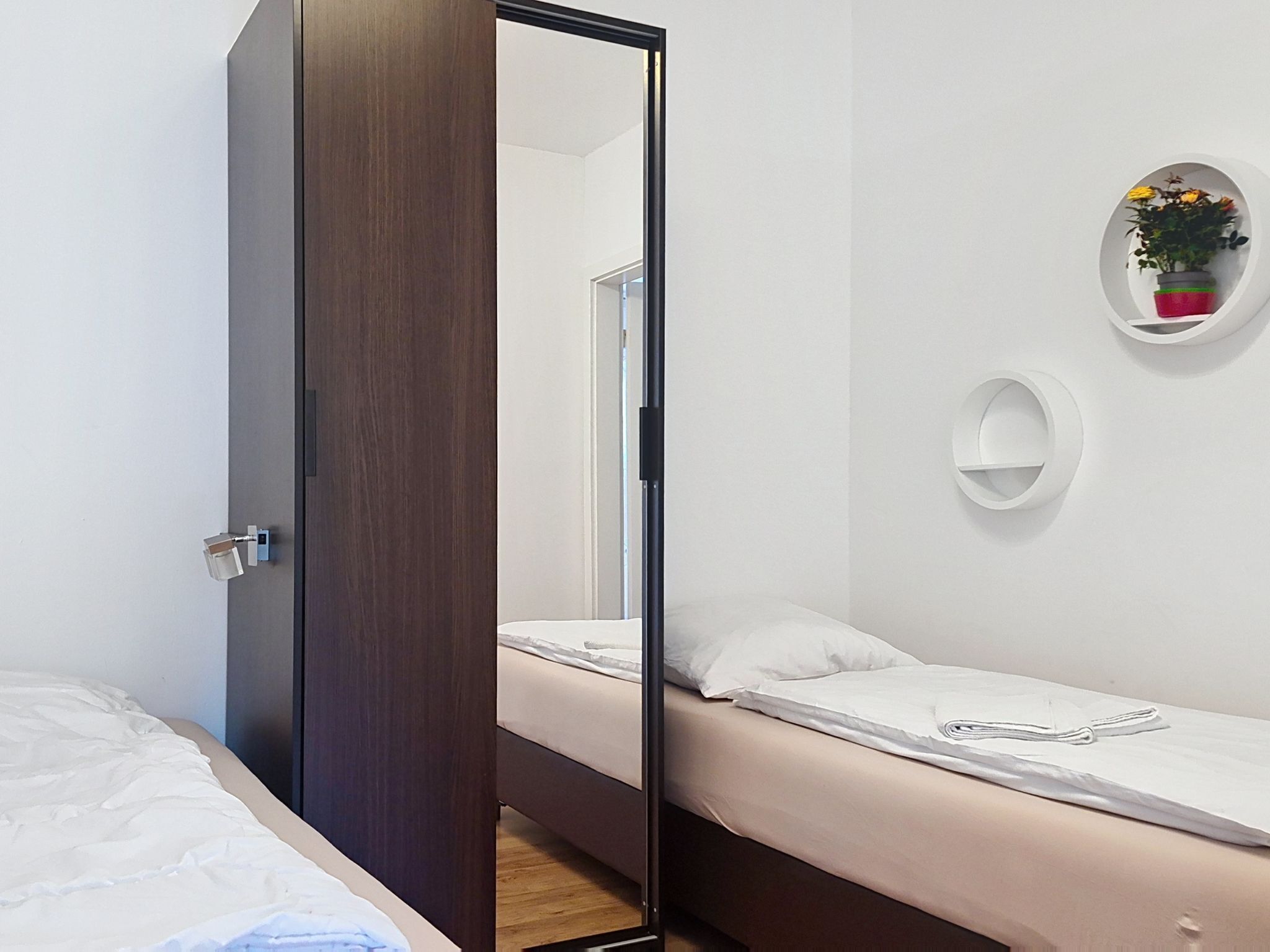 Una habitación con una cama, una mesita de noche, un espejo, un estante en la pared con una planta y una puerta.