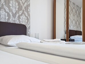 Un lit bien fait, avec du linge de lit blanc et des oreillers, une table de chevet avec une lampe, et un mur recouvert d’un papier peint à motifs.
