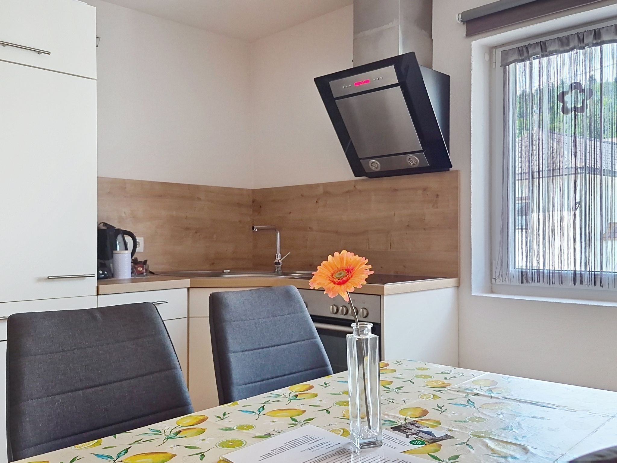 A compact kitchen with a built-in oven, a sink, and a window with blinds. There is a dining table with chairs and a vase with a flower.