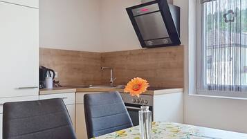 A compact kitchen with a built-in oven, a sink, and a window with blinds. There is a dining table with chairs and a vase with a flower.