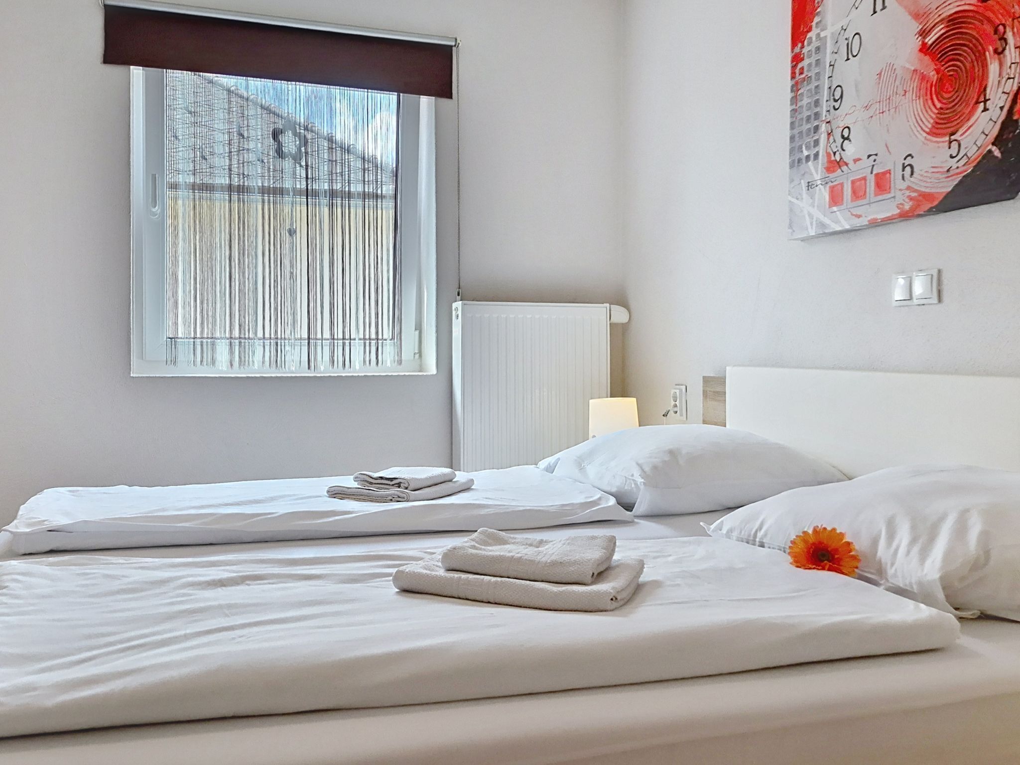Een hotelkamer met een bed, een raam met zonwering, een radiator en een wandklok.