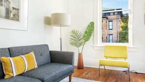 Una sala de estar con un sofá gris, un sillón amarillo, una lámpara de pie, una planta en maceta y una ventana con vistas a edificios.