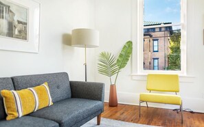 A living room with a grey sofa, a yellow chair, a floor lamp, a potted plant, and a window with a view of buildings. - Park House Carriage, Just Steps from Forsyth Park (Savannah)