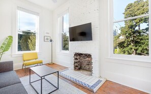 A modern living room with a fireplace, a flat-screen TV mounted on the wall, and large windows offering a view of the outdoors. - Park House Carriage, Just Steps from Forsyth Park (Savannah)