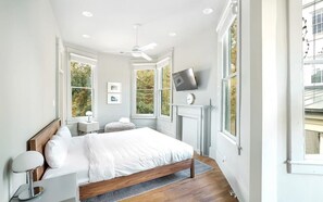 A bedroom with a bed, bedside table, lamp, TV, fireplace, and large windows. - Park House Carriage, Just Steps from Forsyth Park (Savannah)