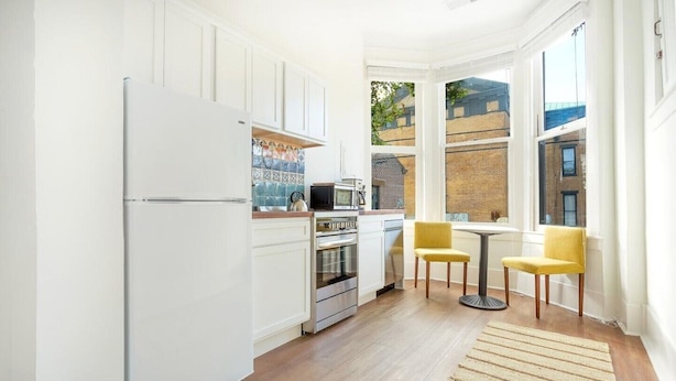 Cocina compacta con gabinetes blancos, refrigerador, horno y microondas. Hay un pequeño comedor con una mesa y dos sillas amarillas.