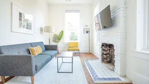 Una sala de estar con un sofá gris, un sillón amarillo, una mesa de centro, una chimenea y un televisor montado en la pared.